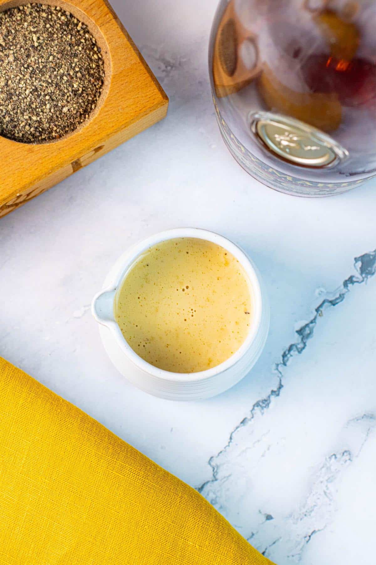 Creamy peppercorn sauce in a small ribbed white jug with a pot of crushed peppercorns and bottle of brandy in the background.