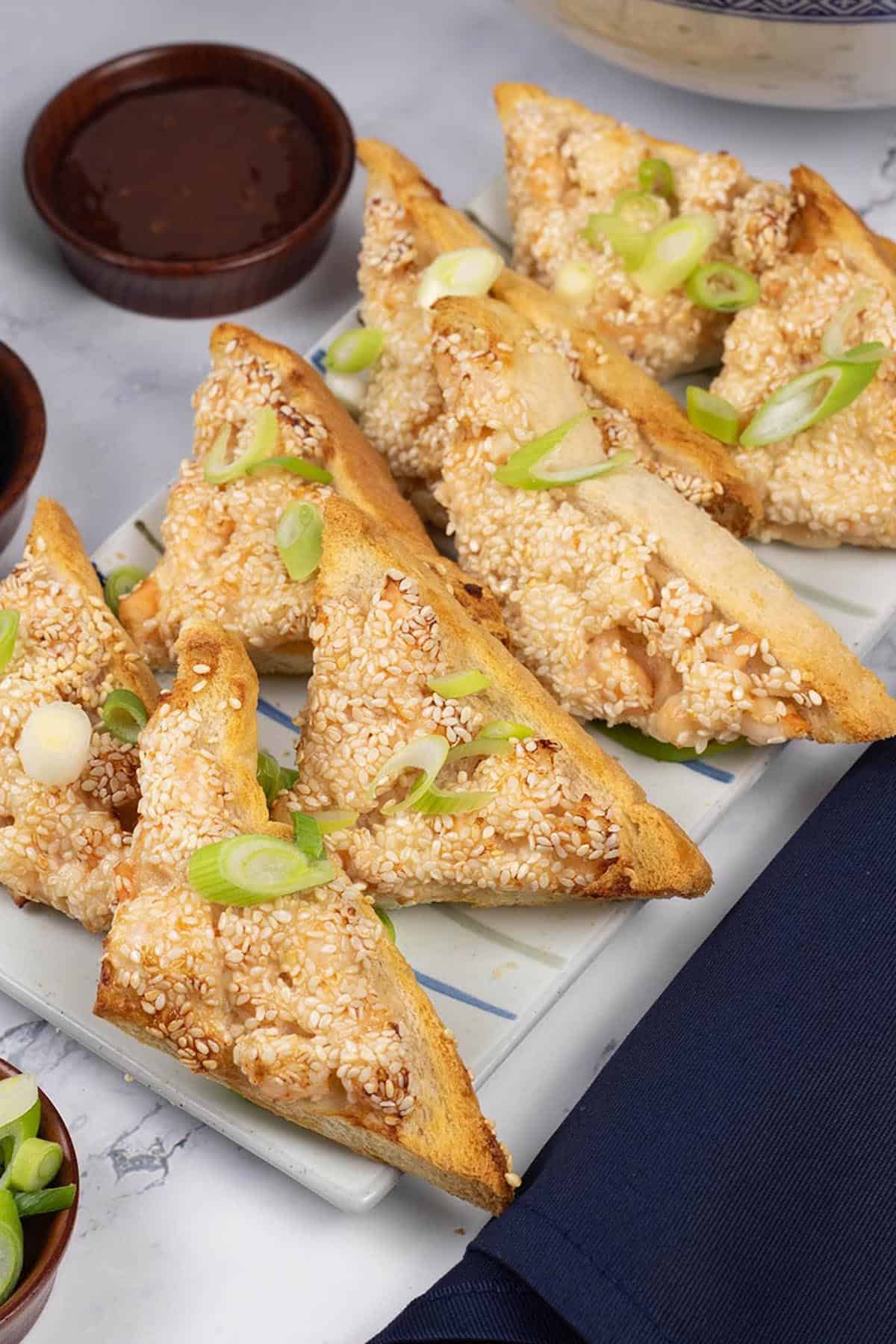 Two portions of easy air fryer prawn toast on a striped platter with small dishes of dipping sauces and spring onions.