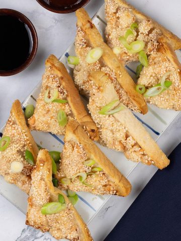 Two portions of easy air fryer prawn toast on a striped platter with small dishes of dipping sauces and spring onions.
