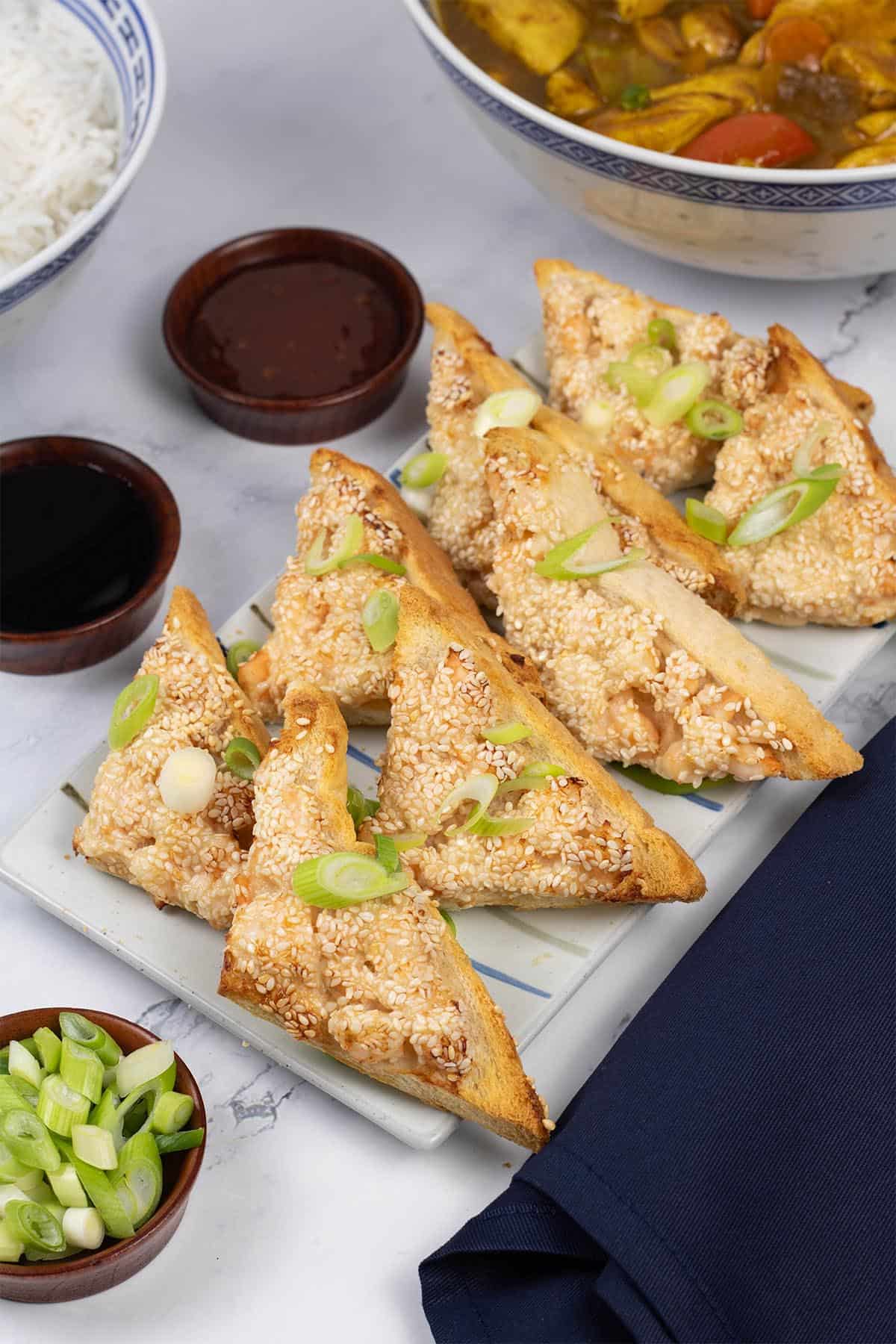 Two portions of easy air fryer prawn toast on a striped platter with small dishes of dipping sauces and spring onions.