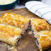 Salmon en croute on a board cut open with a teal and white striped towel and bowls of mashed potatoes and green veg on the side.