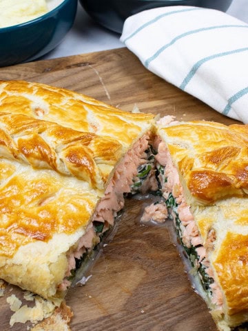 Salmon en croute on a board cut open with a teal and white striped towel and bowls of mashed potatoes and green veg on the side.