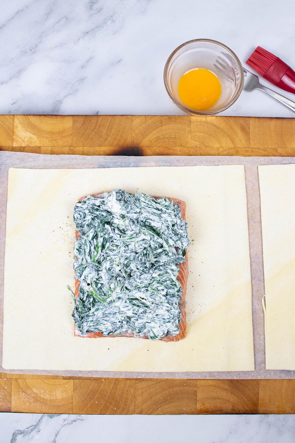 Salmon en croute on a board showing spinach filling, before being cooked, with a fork and bowl of egg yolk and pastry brush on the side.