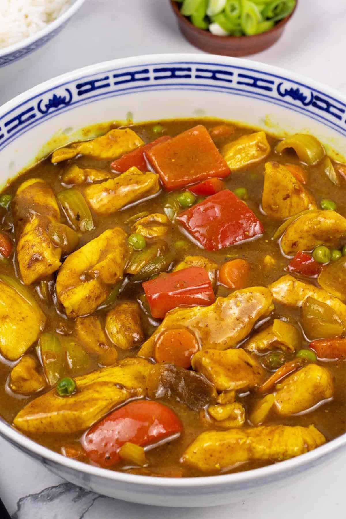 Fakeaway Chinese chicken curry in a large blue and white oriental serving dish with a bowl of rice and small dish of spring onions on the side.