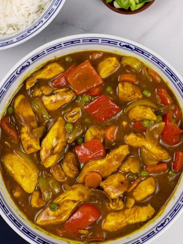 Fakeaway Chinese chicken curry in a large blue and white oriental serving dish with a bowl of rice and small dish of spring onions on the side.