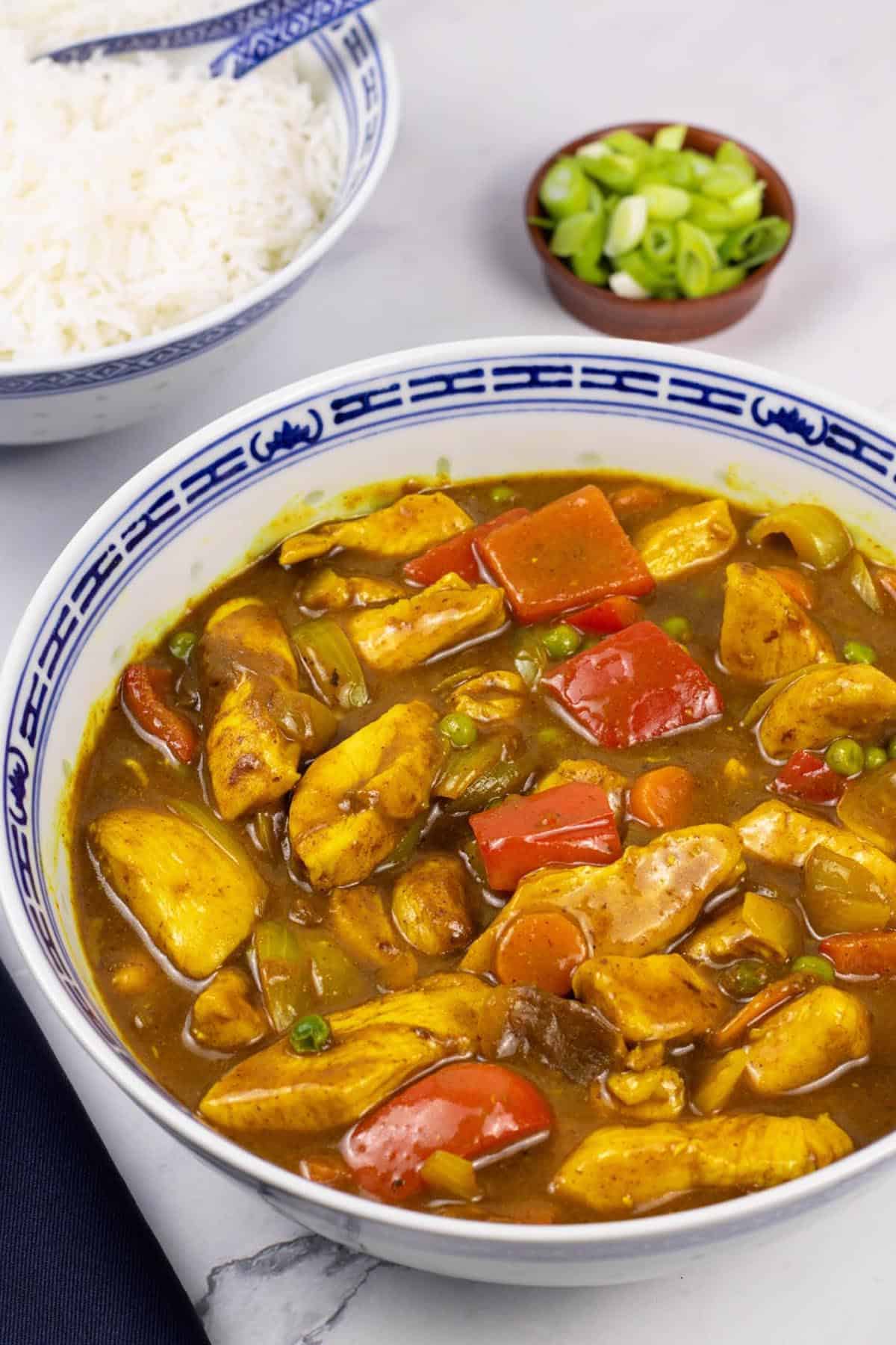 Fakeaway Chinese chicken curry in a large blue and white oriental serving dish with a bowl of rice and small dish of spring onions on the side.