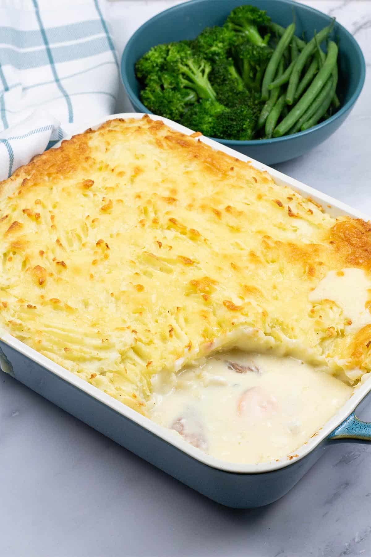 Fish pie in a rectangular ovenproof dish with a bowl of broccoli and green beans, and a teal and white checked towel on the side.