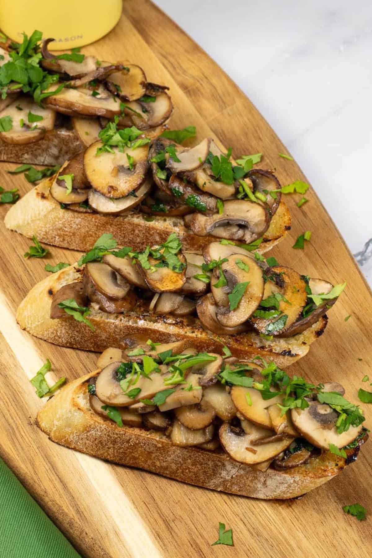 Two portions of garlic mushroom bruschetta on a board with yellow salt and pepper mills and a green napkin on the side.