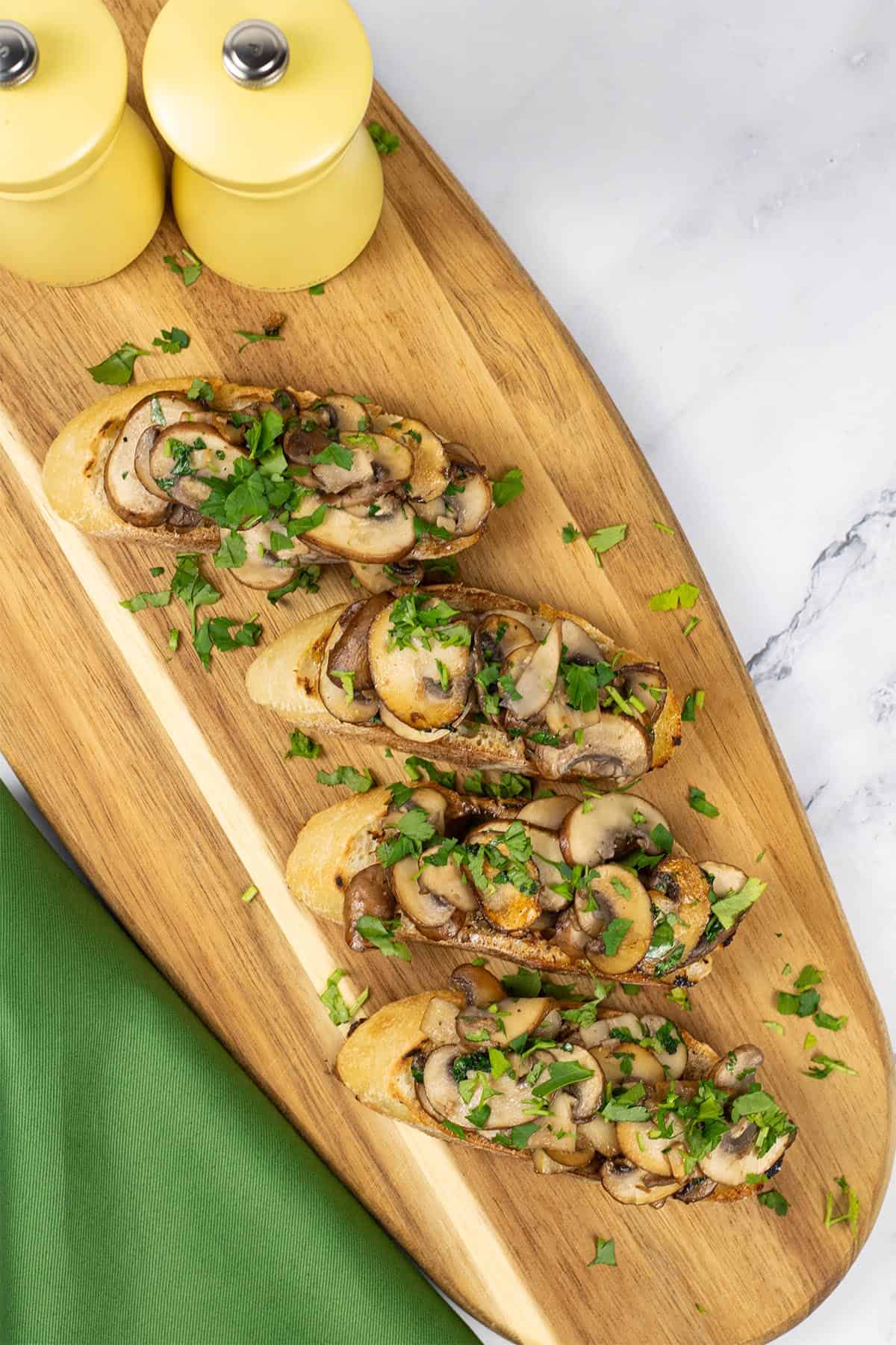 Two portions of garlic mushroom bruschetta on a board with yellow salt and pepper mills and a green napkin on the side.