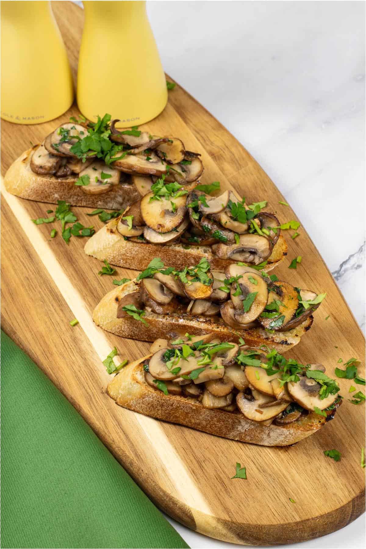 Two portions of garlic mushroom bruschetta on a board with yellow salt and pepper mills and a green napkin on the side.