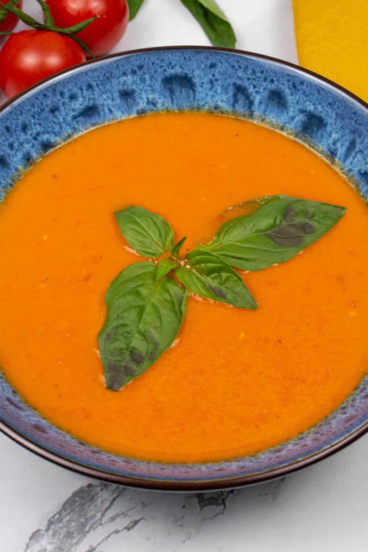 Roast tomato, chilli and fennel soup in a blue patterned bowl, garnished with basil leaves. A bunch of vine tomatoes, basil and a mustard napkin on the side.