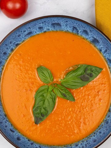 Roast tomato, chilli and fennel soup in a blue patterned bowl, garnished with basil leaves. A bunch of vine tomatoes, basil and a mustard napkin on the side.