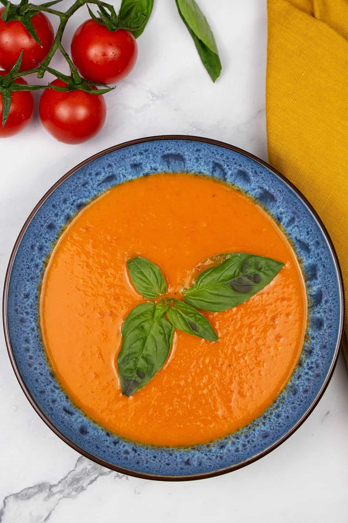 Roast tomato, chilli and fennel soup in a blue patterned bowl, garnished with basil leaves. A bunch of vine tomatoes, basil and a mustard napkin on the side.