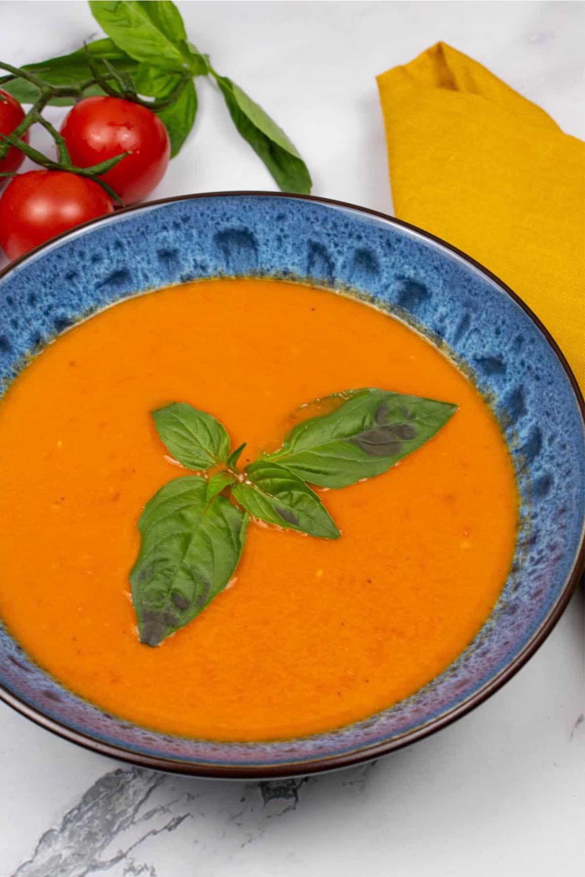 Roast tomato, chilli and fennel soup in a blue patterned bowl, garnished with basil leaves. A bunch of vine tomatoes, basil and a mustard napkin on the side.