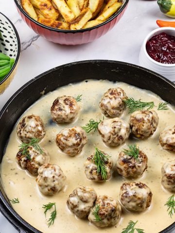 Scandi-style meatballs with gravy in a skillet, with dishes of chips, green beans and lingonberry spread on the side.
