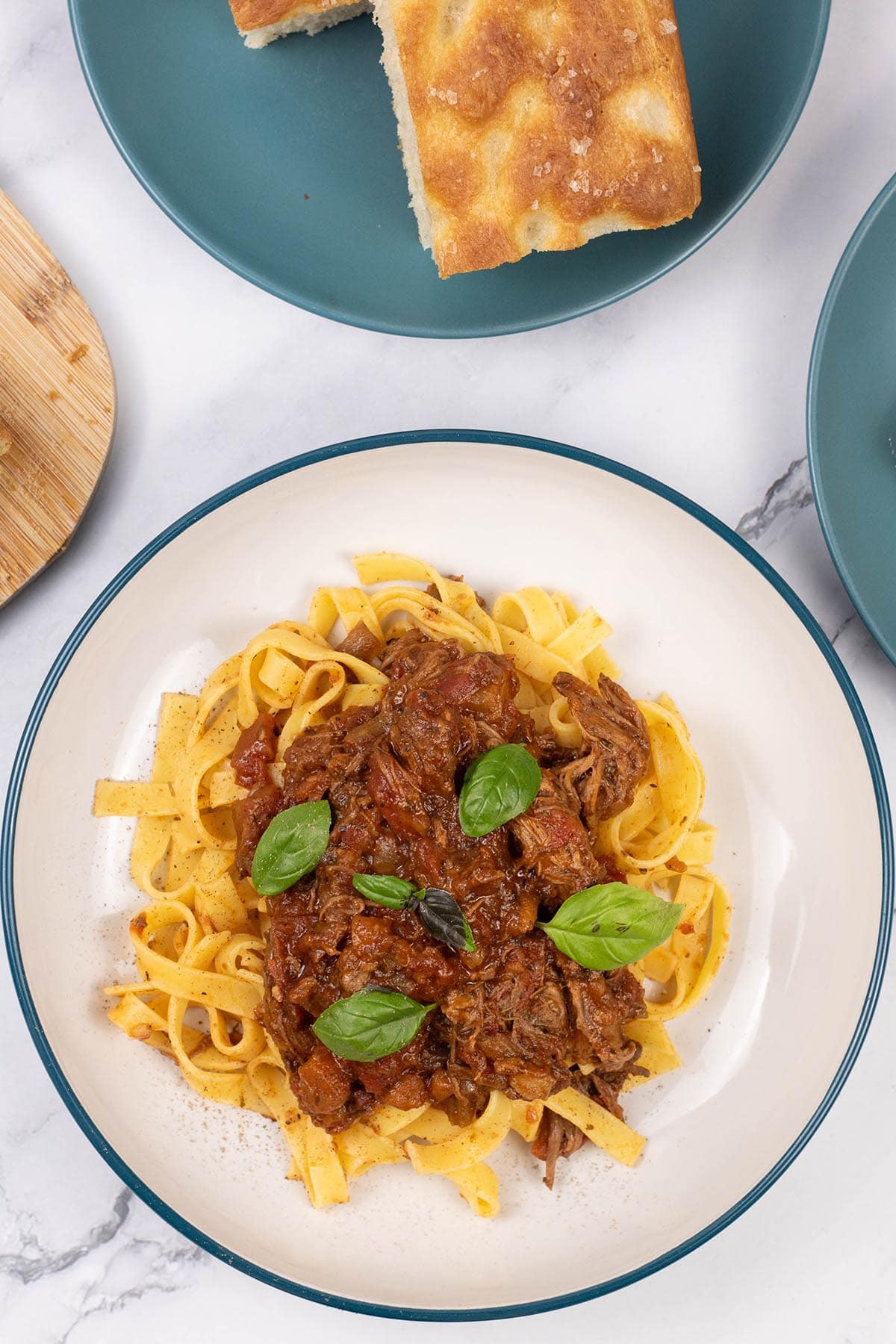 A portion of slow cooker beef ragu in a pasta bowl with tagliatelle and a teal side plate with slices of focaccia in the background.