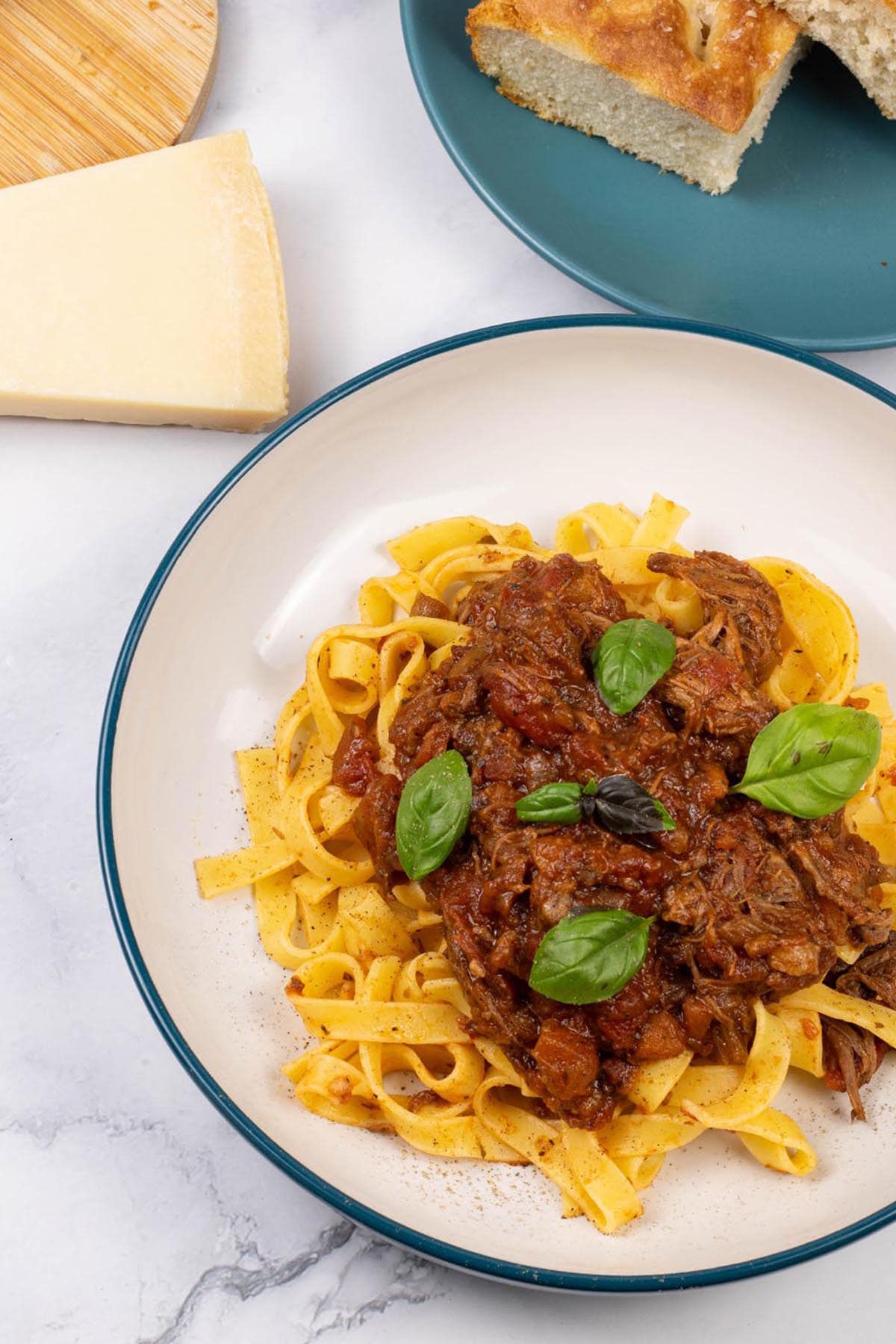 A portion of slow cooker beef ragu in a pasta bowl with tagliatelle and a teal side plate with slices of focaccia in the background.