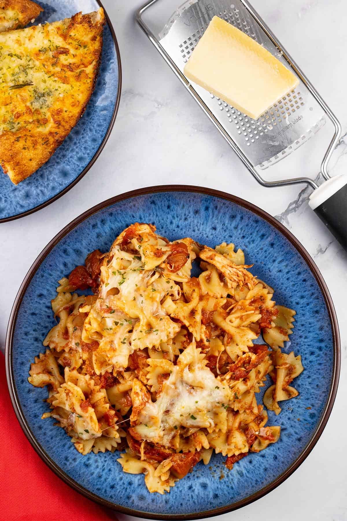A portion of slow cooker chicken parmesan pasta in a blue patterned bowl with a plate of garlic bread, block of parmesan cheese and a red napkin on the side.