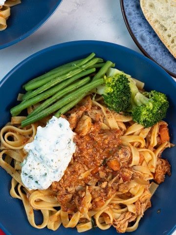 Two portions of slow cooker chicken ragu with basil ricotta in blue bowls, with a bowl of basil ricotta and plate of sliced crusty bread on the side.