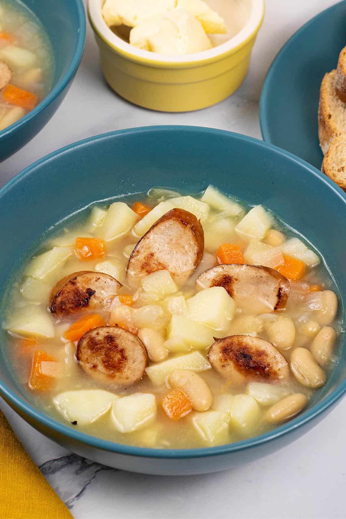 Two portions of smoked sausage, bean and root vegetable soup in teal bowls with a plate of bread and dish of butter on the side.