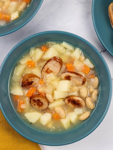Two portions of smoked sausage, bean and root vegetable soup in teal bowls with a plate of bread on the side.