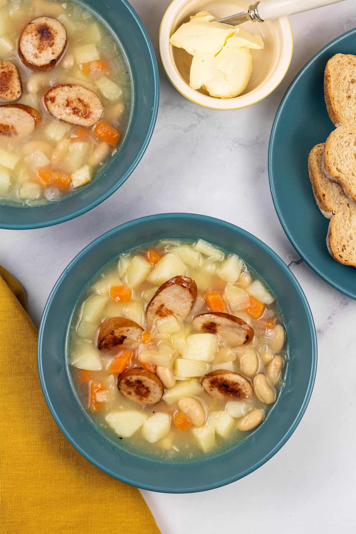 Two portions of smoked sausage, bean and root vegetable soup in teal bowls with a plate of bread and dish of butter on the side.