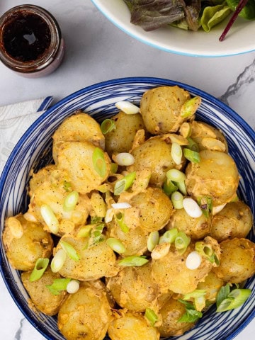Smoky chipotle potato salad in a blue and white serving bowl, with a jar of chipotle paste and bowl of salad leaves on the side.