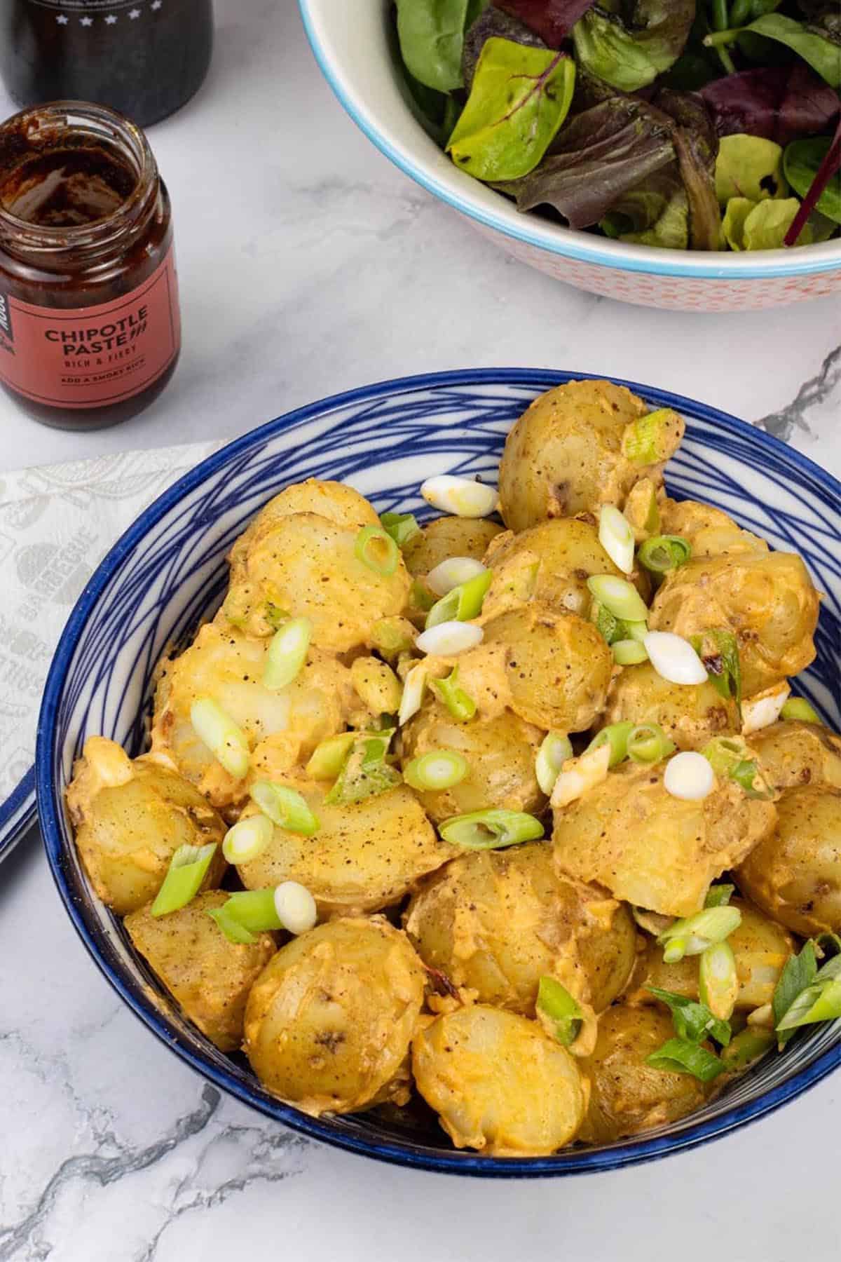 Smoky chipotle potato salad in a blue and white serving bowl, with a jar of chipotle paste and bowl of salad leaves on the side.