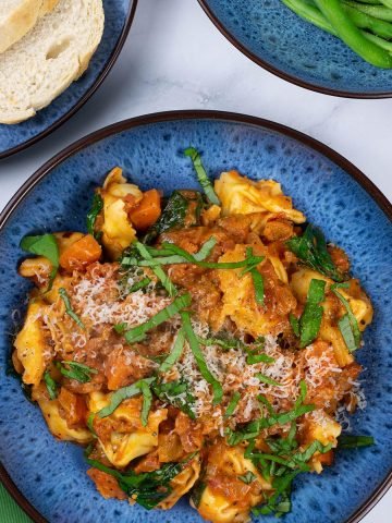 A portion of tortellini with creamy tomato and spinach sauce in a blue patterned bowl with a bowl of green beans and plate of sliced bread on the side.