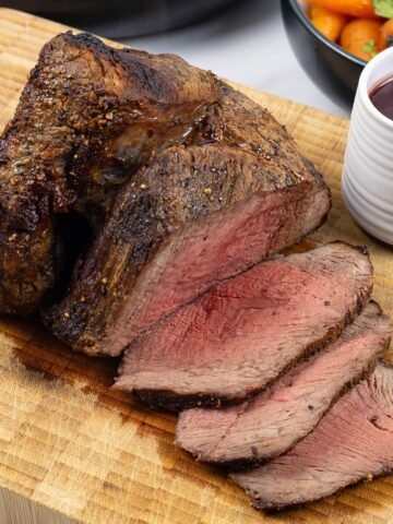 Air fryer roast beef sitting on a board with 3 slices carved, a small jug of red wine jus and a bowl of chantenay carrots on the side.