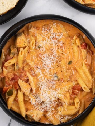 Two portions of cajun chicken and chorizo pasta in black bowls with a plate of sliced baguette on the side.