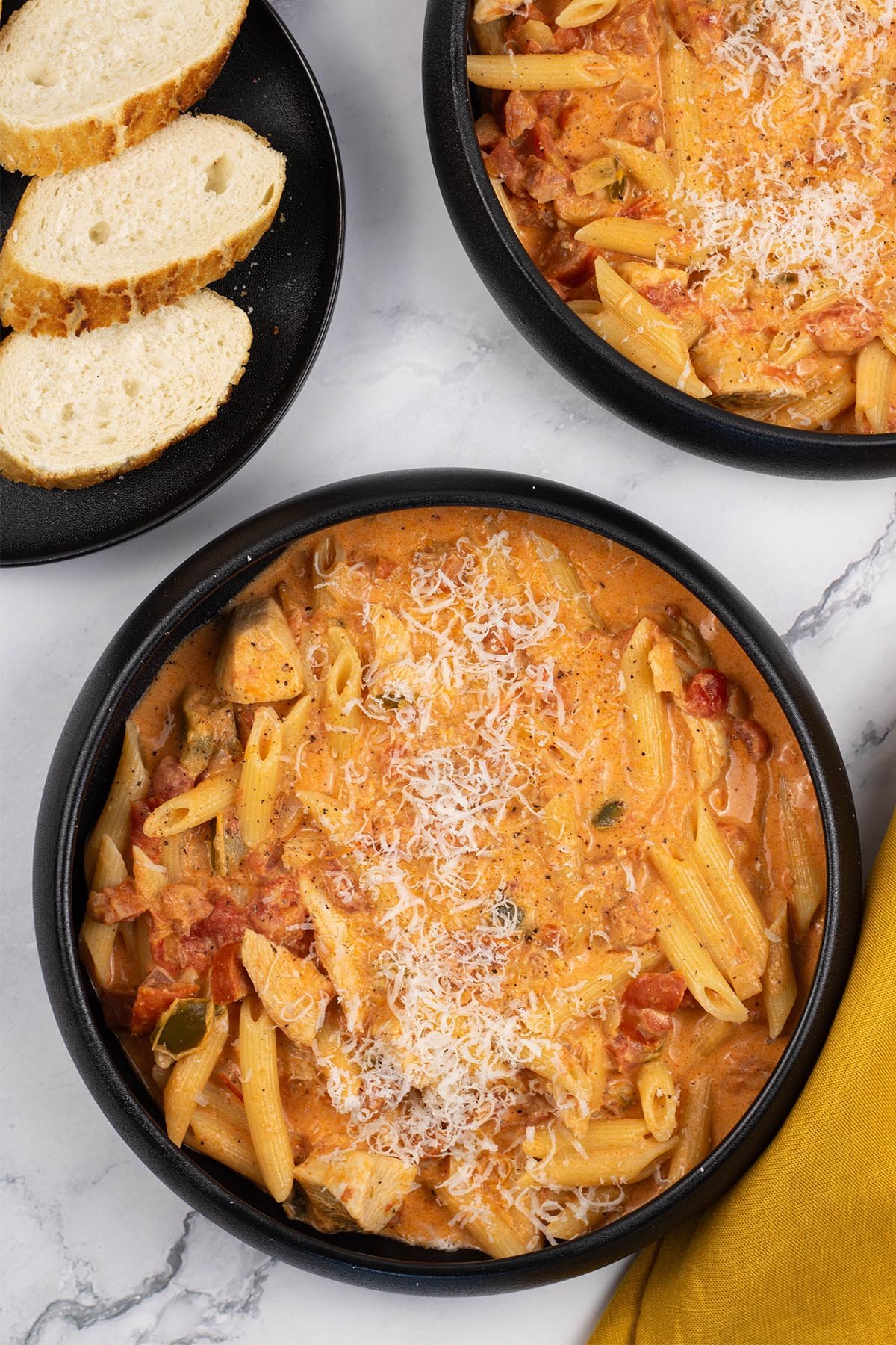 Two portions of cajun chicken and chorizo pasta in black bowls with a plate of sliced baguette on the side.