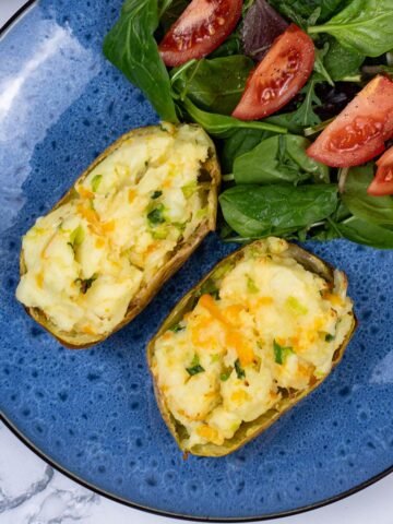 Cheese and spring onion baked potato on a blue patterned dinner plate with salad leaves and tomato wedges on the side.