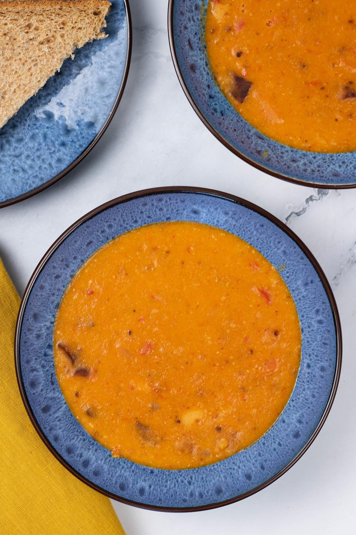 Two portions of chorizo and lentil soup in blue patterned bowls with a plate of sliced brown bread on the side.
