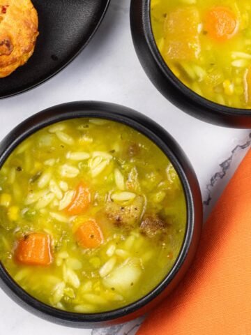 Two portions of chunky mixed vegetable soup made in the Thermomix, in black bowls with a plate of chorizo, sweetcorn and chilli muffins on the side.