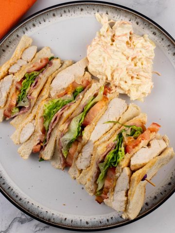 A club sandwich and coleslaw on a black and white patterned dinner plate with an orange napkin on the side.