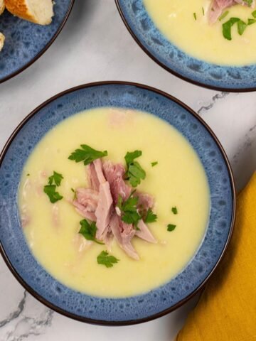 Two portions of creamy leek and potato soup with ham in blue patterned bowls, with a plate of sliced baguette on the side.