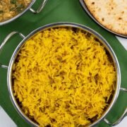 Golden pilau rice in a balti dish with a dish of slow cooker chicken and potato curry, and plate of chapatis in the background.