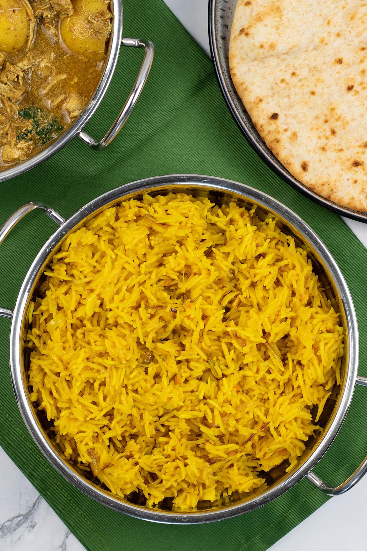 Golden pilau rice in a balti dish with a dish of slow cooker chicken and potato curry, and plate of chapatis in the background.