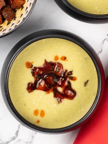Two portions of leek, butter bean and crispy chorizo soup in black bowls with a bowl of crispy chorizo on the side.