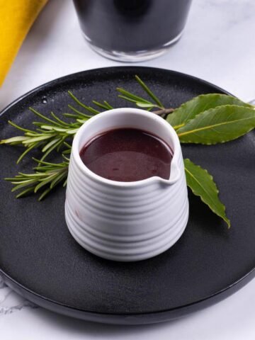 Red wine jus in a small white ribbed jug sitting on a black side plate decorated with 2 sprigs of rosemary and 3 bay leaves.