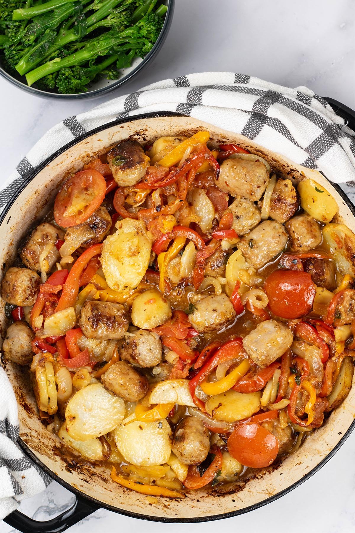 Salsiccia con patate e pomodori al forno in a large round casserole with a bowl of broccoli and green beans on the side.