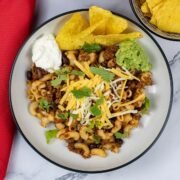 A portion of slow cooker cheesy chilli mac in a black and cream bowl garnished with sour cream, tortilla chips and guacamole.