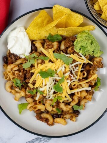 A portion of slow cooker cheesy chilli mac in a black and cream bowl garnished with sour cream, tortilla chips and guacamole.
