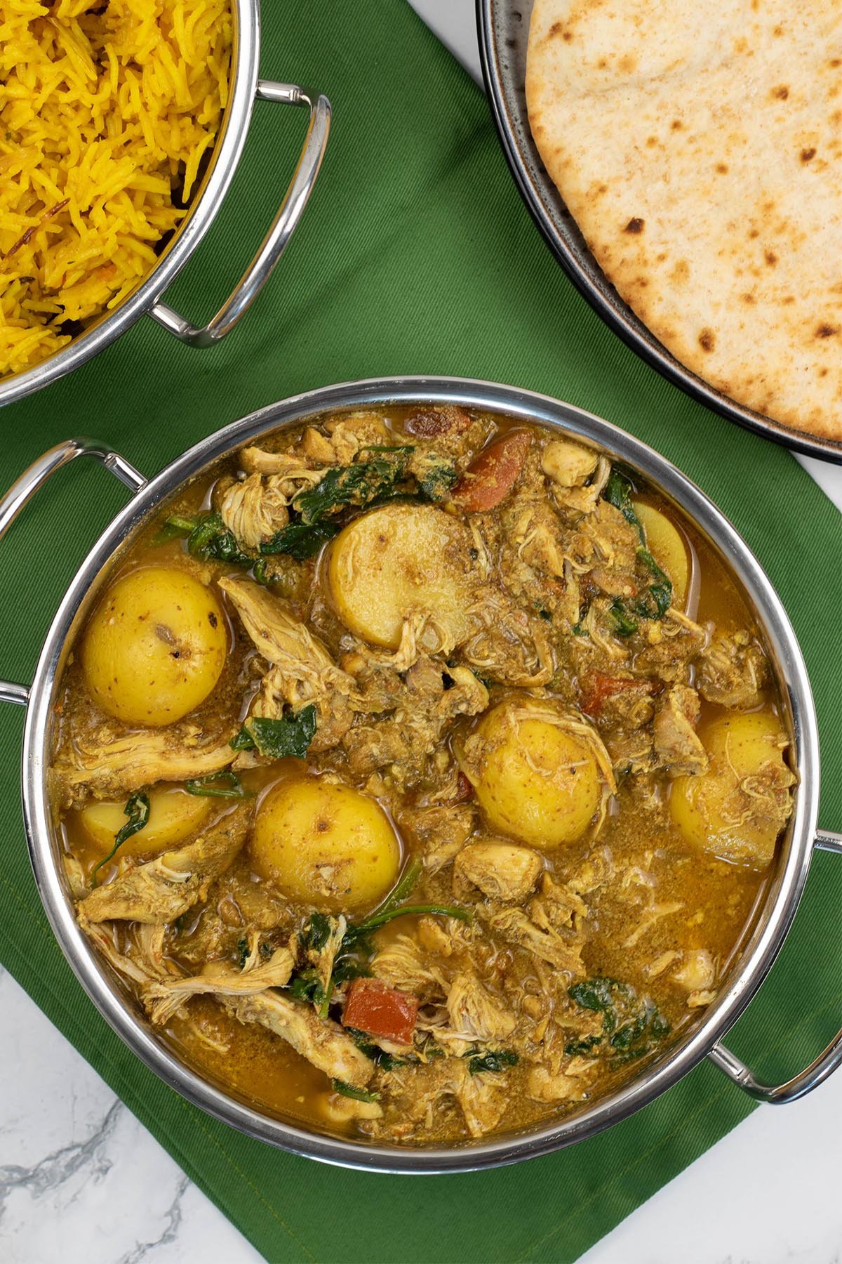 Slow cooker chicken and potato curry in a balti dish with golden pilau rice and chapatis in the background.