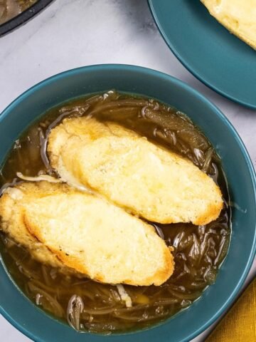 A portion of french onion soup in a teal bowl with 2 slices of baguette covered with melted cheese.