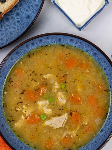 A portion of slow cooker roast chicken soup in a blue patterned bowl with a plate of sliced Portuguese water bread and dish of garlic and lemon yogurt on the side.
