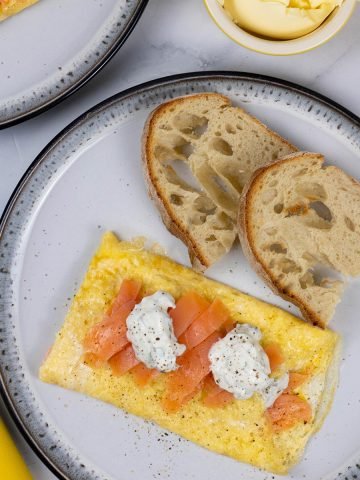 A portion of smoked salmon and lemon herb cream cheese omelette on a black and white patterned dinner plate with a slice of toasted Portuguese water bread.