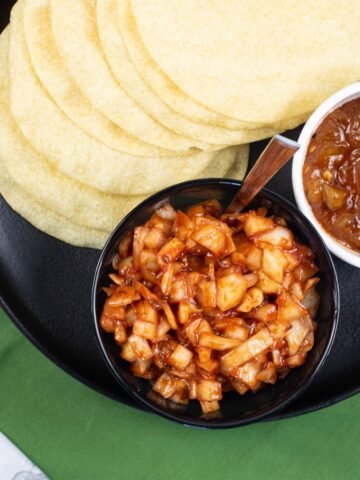 Spicy onions in a black serving bowl sitting on a black dinner plate with poppadoms and a dish of mango chutney.