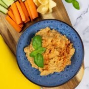 Sun-dried tomato and basil houmous in a blue patterned serving bowl sitting on a board with carrot and cucumber sticks and lentil crisps.